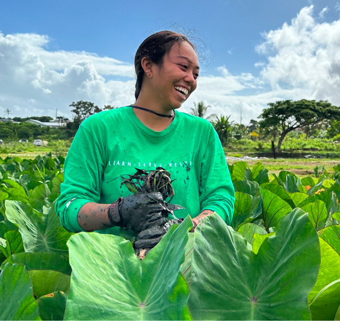 A person smiling while holding plants in a lush green field.