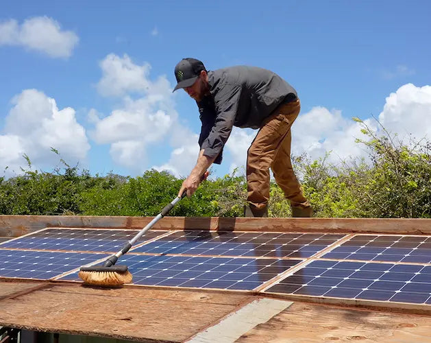 Person cleaning solar panels on a roof with a long brush.